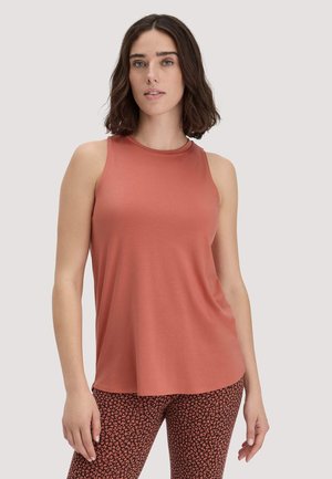 Woman with shoulder-length brown hair wearing a sleeveless rust-colored top and patterned brown and orange pants, standing against a neutral background.