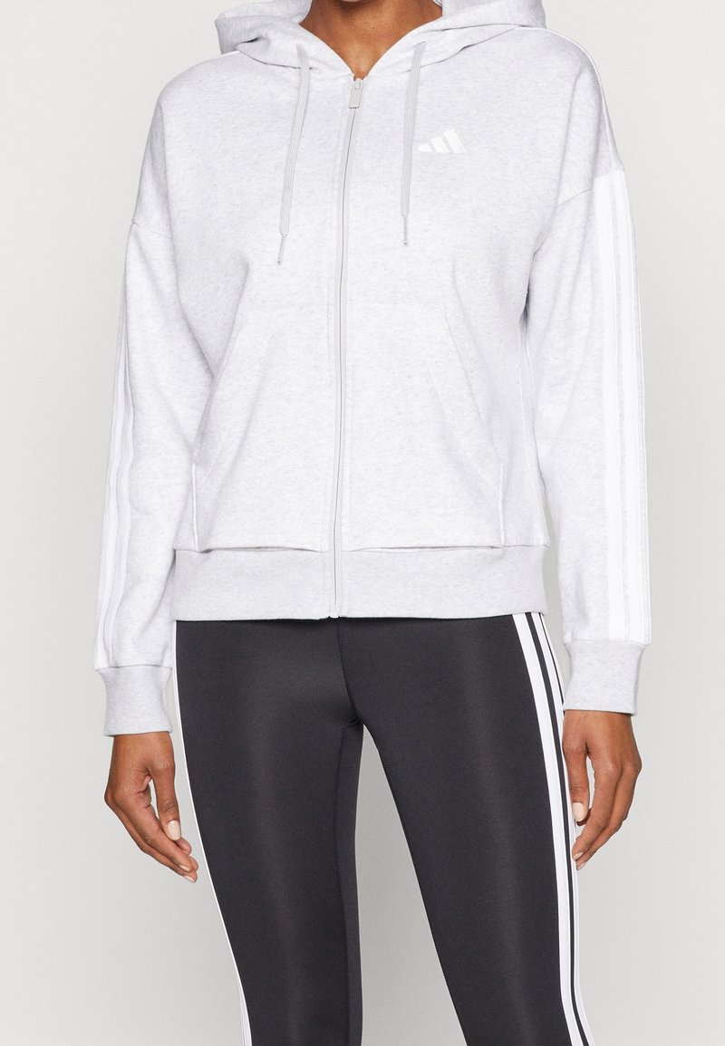 Grå zip hoodie i mjukt material, med huva, två framfickor och vita randiga detaljer längs ärmarna. Vit logotyp på bröstet.