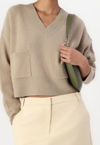Beżowy sweter dzianinowy z dekoltem w serek i dwoma przednimi kieszeniami, połączony z kremowymi spodniami oraz zieloną torebką na pasek.