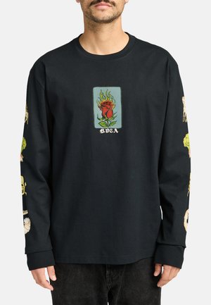 Mand iført sort langærmet skjorte med flammende rose grafik og tekst på brystet samt forskellige farverige print på begge ærmer.