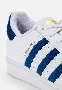 White sneaker with blue textured suede stripes, lace-up design, gold "SUPERSTAR" lettering, and a rubber sole.