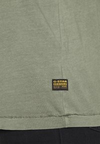 Olivgrön bomullströja med rak skärning, som har en svart etikett med gul text nära fållen, som visar varumärke och skötselråd.