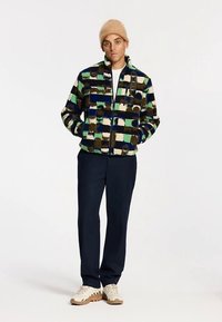 Többszínű polárdzseki zöld, tengerészkék és krémszínű négyzetekkel, cipzáras záródással és oldalzsebekkel; tengerészkék nadrággal és bézs színű lábbelivel párosítva.