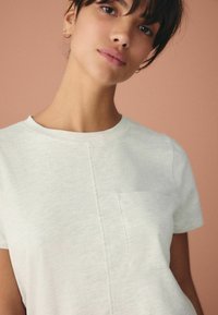 Hellgraues T-Shirt aus weichem Stoff, mit Rundhalsausschnitt und kleiner Brusttasche. Das Design hat subtile Textur- und Nähdetails.