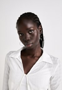 Young woman with braided hair wearing hoop earrings and a white collared shirt, looking directly at the camera against a plain background.
