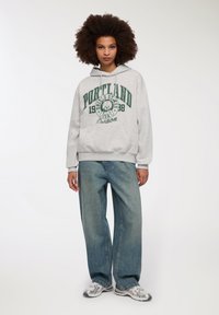 Felpa grigia con grafica "PORTLAND 1988" in verde, abbinata a jeans a gamba larga blu chiaro e sneakers argento. Tessuto liscio e vestibilità rilassata.