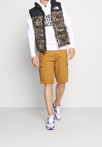 Uomo in felpa bianca con gilet imbottito a fantasia mimetica nera e verde, pantaloni cargo giallo senape e sneaker bianche con accenti viola, in piedi su un pavimento grigio.
