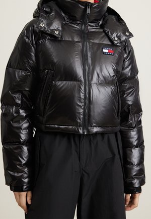 Black shiny puffer jacket with high collar and Tommy Jeans logo, worn with black pants, showing front view from shoulders to hips.