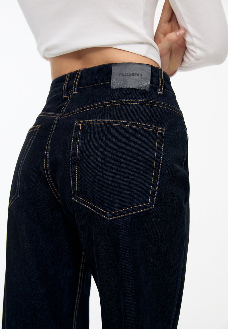 Dunkelblaue Jeans aus Denim mit orangefarbenen Nähten, ausgestattet mit einer Gesäßtasche und einem grauen Etikett mit "PULL&BEAR" am Bund.