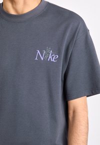 T-shirt de algodão cinza escuro com um logo "Nike" bordado em lavanda no lado esquerdo do peito. Mangas curtas e design de gola redonda padrão.