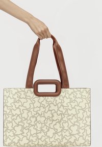 Bolso tote crema con una textura estampada, asas de tela marrón y un acento rectangular marrón en la parte superior.