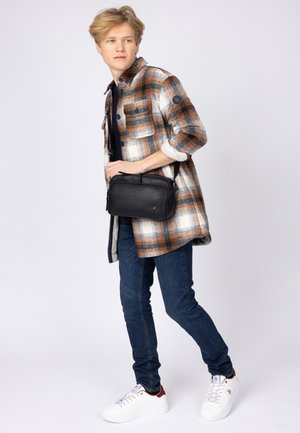 Svart läder crossbody-väska med dragkedjestängning och justerbar rem, med en liten logotyp på framsidan. Bärs av en modell i en rutmönstrad skjorta.