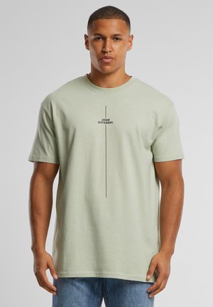 Vyras, dėvintis šviesiai žalią marškinėlį su užrašu „THINK DIFFERENT.“ ir vertikaliu juodu dryžu priekyje, derinamas su mėlynais džinsais.