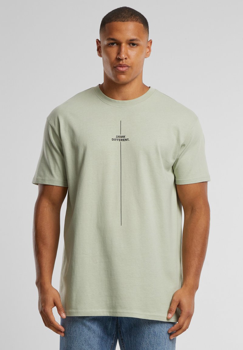 Mann trägt ein hellgrünes T-Shirt mit dem Text „THINK DIFFERENT.“ und einer schwarzen vertikalen Linie vorne, kombiniert mit blauen Jeans.