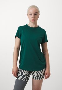 Zelena majica kratkih rukava s rebrastom teksturom, u kombinaciji s kratkim hlačama u crno-bijelom zebrastrukom uzorku. Manekenka prikazuje protezu za nogu.