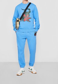 Persona vestida con sudadera azul y joggers con estampados de cocodrilo verde y flores rojas, zapatillas blancas y un bolso bandolera beige.