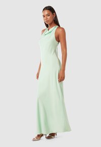 Cowl-neck, sleeveless maxi dress in light mint green fabric. Smooth texture, form-fitting design, and side slits. Worn with silver heels.