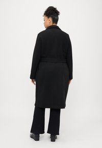 Musta pitkä takki vyöllä, leikkauskauluksella ja sileällä pinnalla. Yllä mustat leveälahkeiset housut ja tukevakorkoiset kengät.