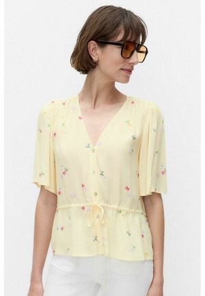 Kobieta o krótkich brązowych włosach, ubrana w żółtą kwiecistą bluzkę z zapięciem na guziki z przodu, ze sznurkiem w talii i dużymi ciemnymi okularami przeciwsłonecznymi, patrząca w prawo.