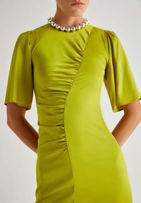 Abito verde lime realizzato in tessuto morbido, con un design arricciato su un lato, maniche a sbuffo corte e una collana di perline argento.