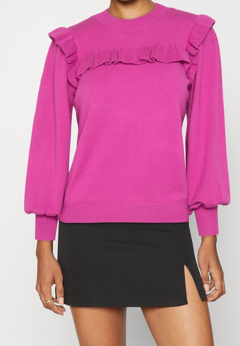 Femme portant un pull à manches longues rose vif avec des volants sur les épaules et la poitrine, associé à une mini-jupe noire fendue sur le côté.