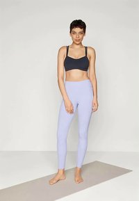 Mladá žena stojí bosá na jogové podložce, oblečená v černém sportovním brá a světle fialových legínách na jednobarevném pozadí.