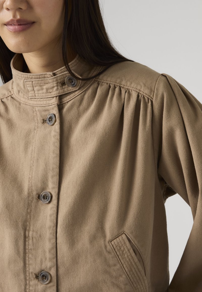 Mujer con chaqueta beige abotonada, con cuello alto y detalle fruncido en el hombro, sobre un fondo liso.