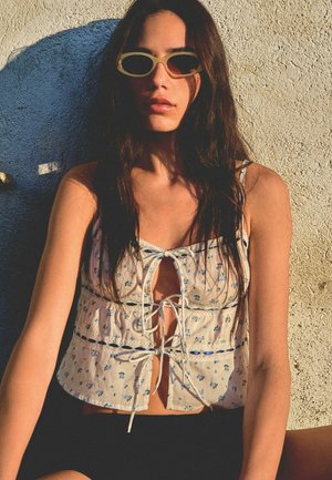Hvid blomsteret crop top med blonde detaljer, der har en lukning foran. Modellen bærer lyse ovale solbriller. Baggrunden er struktureret beige.