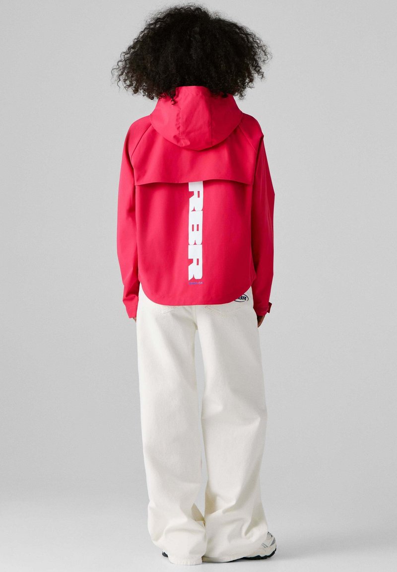 Person med krøllet hår iført en rød hættejakke med hvid lodret tekst og løse hvide bukser, stående mod en grå baggrund.