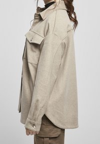Beige uldjakke med en afslappet pasform, sidelommer og knapdetaljer på ærmerne, vist på en model fra siden.
