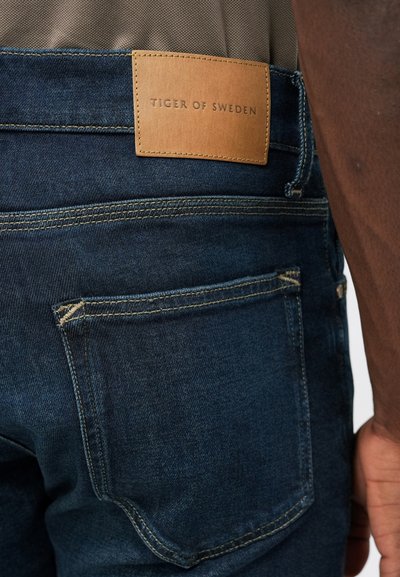 Sötét farmernadrág barna bőr folttal, amelyen a "Tiger of Sweden" felirat látható, látható varrással és a hátsó zseb részleteivel.