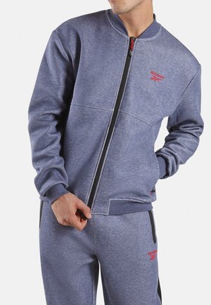 Man draagt een blauw Reebok trainingspak met rood logo, ritst het jack dicht, hand in de zak, bijgesneden op kin en knieën.