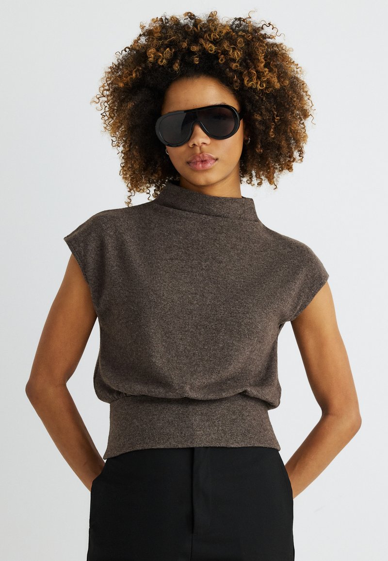 Lokkis juustega naine, kes kannab suuri musti päikeseprille, pruunikat varrukateta kõrge kaelusega topi ja musti pükse, taustal lihtne hele taust.