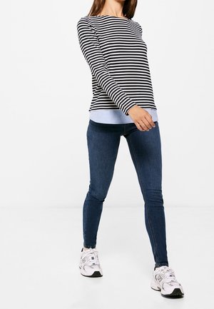 Frau trägt ein schwarz-weiß gestreiftes Langarmshirt, blaue enge Jeans und weiße Turnschuhe, steht vor einem weißen Hintergrund.
