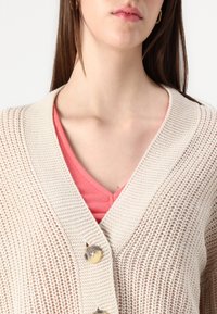 Lys beige strikket cardigan med V-hals, med to store knapper. Brukt over en korallfarget T-skjorte, som viser et mønstret stoff.