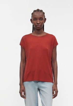 Vrouw met gevlochten haar die een effen roestkleurig T-shirt met korte mouwen en lichtblauwe spijkerbroek draagt, staand tegen een witte achtergrond.