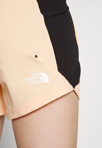 Acercamiento a unos pantalones cortos deportivos de melocotón claro y negro con el logo de The North Face en el muslo izquierdo, llevados por una persona.