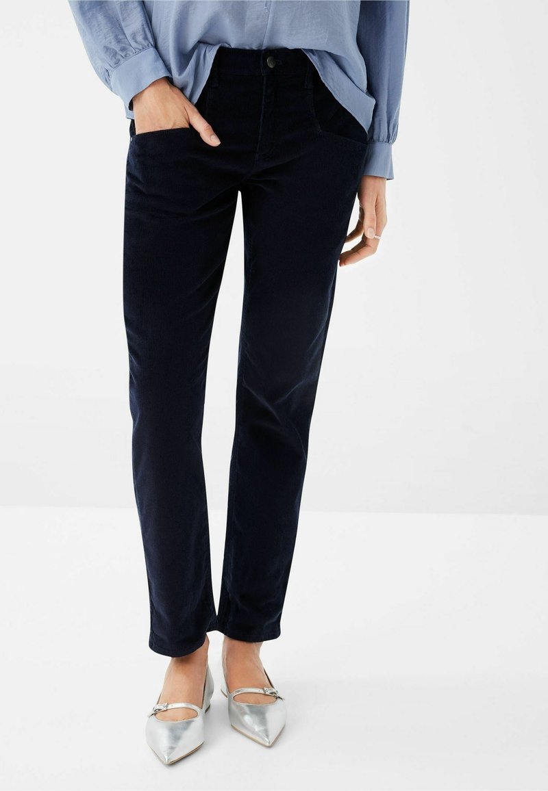 Femme portant un pantalon bleu foncé, une chemise à manches longues bleu clair et des chaussures argentées à bout pointu, debout avec une main dans la poche.