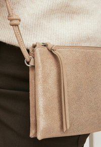 Bežová trblietavá crossbody taška s textúrovaným povrchom, so zipsom a odnímateľným popruhom, položená na krémovom svetre a tmavých nohaviciach.