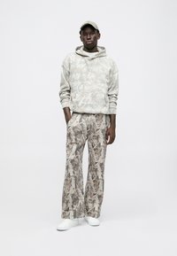 Felpa grigia chiara con motivo, dotata di tasca frontale, abbinata a pantaloni larghi con motivo in tonalità di beige e marrone. Sneakers bianche.