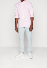 Mann in einem hellrosa, knöpfbaren Hemd mit hochgekrempelten Ärmeln, hellblauen Jeans und weißen Sneakers, der vor einem schlichten Hintergrund steht.