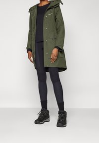 Personne portant un imperméable à capuche vert olive, un haut à manches longues noir, des leggings noirs, des chaussettes noires et des chaussures de randonnée noires, debout devant un fond uni.