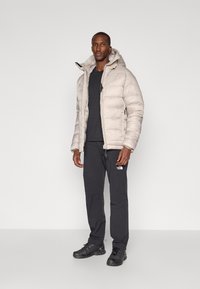 Geacă puffer, bej deschis, cu glugă și închidere cu fermoar. Cămașă neagră cu guler rotund, pantaloni negri și pantofi sport închiși la culoare. Design simplu.