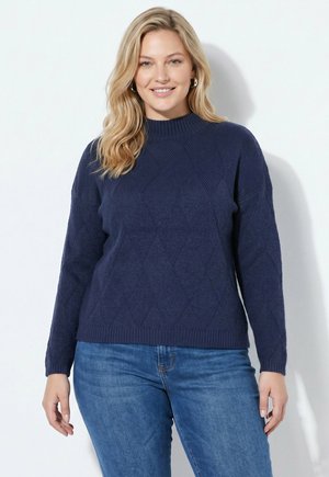 Lachende vrouw met blond haar draagt een marineblauwe gebreide trui en blauwe spijkerbroek, staand tegen een effen witte achtergrond.