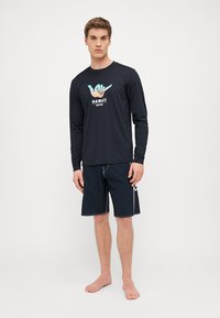 Rash guard z długim rękawem w kolorze granatowym z grafiką "HAWAII" oraz symbolem ręki surferów. W zestawie z ciemnymi szortami na desce z kontrastowym wykończeniem.