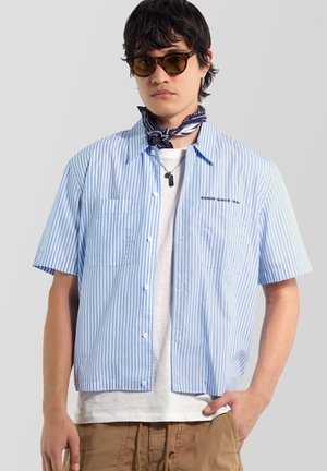 Mann trägt braune Sonnenbrille, blau-weiß gestreiftes Kurzarmhemd über weißem T-Shirt, dunkelblaues Halstuch und braune Hose vor einfarbigem Hintergrund.