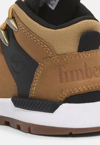 Braune und schwarze Sneaker mit einer strukturierten Oberseite, gebrandetem Logo, metallischen Ösen und einer weißen Gummisohle mit einem gemusterten Profil.
