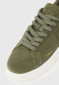 Sapatilhas de camurça verde-azeitona com atacadores planos, sola de borracha branca e marcação em dourado. Parte superior texturizada com detalhes em costura.