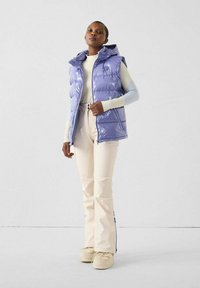 Paarse bodywarmer met capuchon, doorgestikt ontwerp en glanzende afwerking, gecombineerd met lichtbeige broek en beige schoenen.