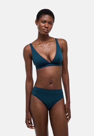 Femme mince portant un maillot de bain deux pièces couleur sarcelle, debout avec une expression neutre sur un fond gris clair uni.